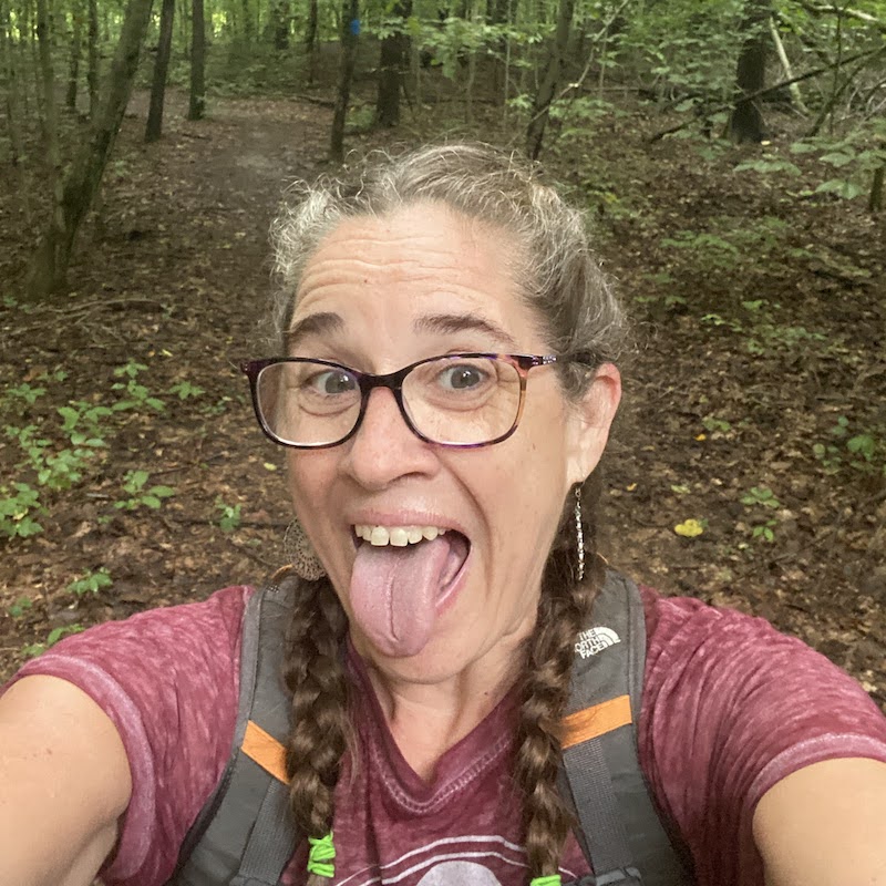 Close-up face selfie of Jolynn walks in a wood with red t-shirt and daypack. She is happy and sticking out her tongue.