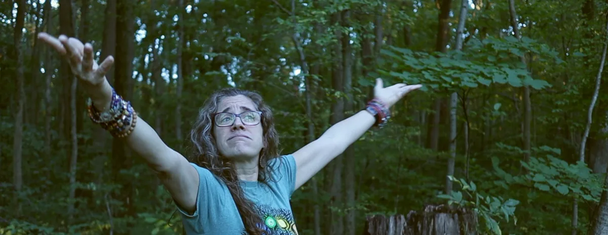 Jolynn standing in the woods with arms raised wide, looking up toward the trees.